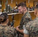 Three military personnel in MARPAT camouflage uniforms are gathered in what appears to be an ordnance or EOD (Explosive Ordnance Disposal) training room or armory. One female service member is holding a dark-colored hand grenade while a male service member facing the camera appears to be examining or discussing an item. The shelves behind them display a large collection of inert training munitions, rockets, and ordnance items.