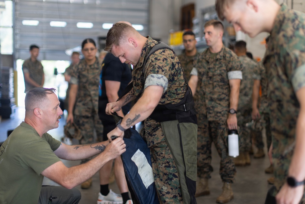 A group of military personnel, appearing to be U.S. Marines based on their MARPAT camouflage uniforms, are gathered inside what appears to be a hangar or warehouse facility. A standing Marine wearing a tactical vest is being assisted or inspected by a seated individual in an olive green t-shirt, who appears to be examining or adjusting gear. Other Marines in uniform stand in line or observe in the background.