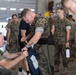 A group of military personnel, appearing to be U.S. Marines based on their MARPAT camouflage uniforms, are gathered inside what appears to be a hangar or warehouse facility. A standing Marine wearing a tactical vest is being assisted or inspected by a seated individual in an olive green t-shirt, who appears to be examining or adjusting gear. Other Marines in uniform stand in line or observe in the background.