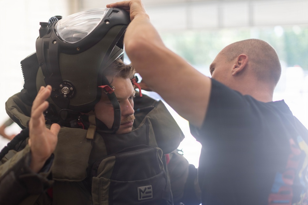 Two individuals are shown in what appears to be a training or equipment fitting scenario. One person is wearing a large protective helmet or flight helmet with a visor, while a second person assists in adjusting or securing the headgear. The setting appears to be an indoor facility, possibly a military or emergency services training environment.