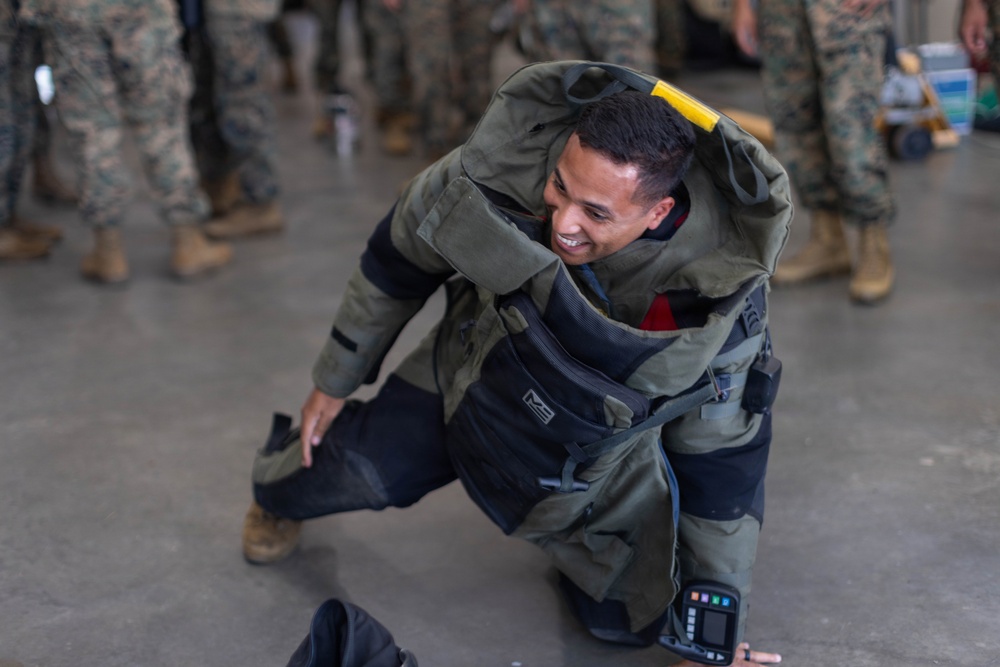 A smiling military service member is kneeling on a concrete floor while donning or demonstrating a bomb disposal (EOD) suit. Other personnel in camouflage uniforms are visible in the background, suggesting a training or demonstration environment. The individual appears relaxed and in good spirits despite wearing the heavy protective equipment.