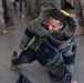 A smiling military service member is kneeling on a concrete floor while donning or demonstrating a bomb disposal (EOD) suit. Other personnel in camouflage uniforms are visible in the background, suggesting a training or demonstration environment. The individual appears relaxed and in good spirits despite wearing the heavy protective equipment.