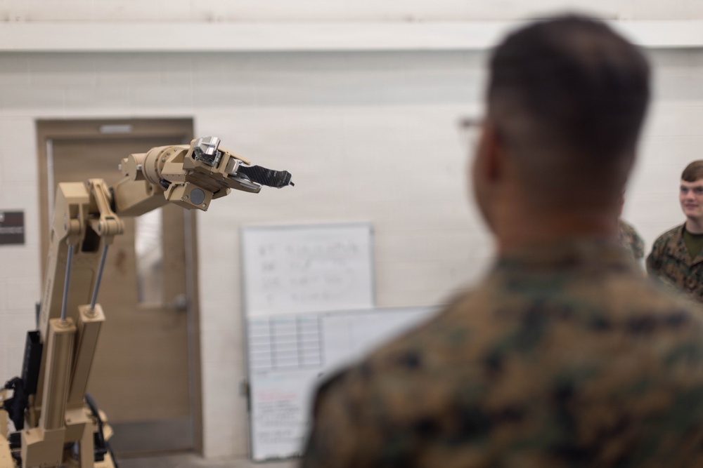 Military personnel in camouflage uniforms are observing a tan/beige robotic arm in what appears to be an indoor facility or training room. The robotic arm is extended and features a gripper/claw end effector. The setting suggests a demonstration or training exercise involving robotic technology.