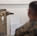 Military personnel in camouflage uniforms are observing a tan/beige robotic arm in what appears to be an indoor facility or training room. The robotic arm is extended and features a gripper/claw end effector. The setting suggests a demonstration or training exercise involving robotic technology.