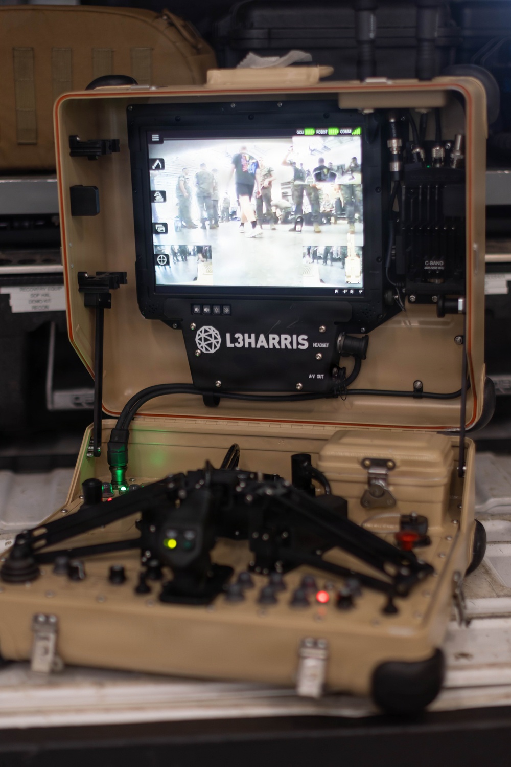 A L3Harris ground control unit (GCU) is shown open in a tan/desert-colored ruggedized carrying case. The system features a monitor displaying a live or recorded camera feed of a group of people, along with a dual-joystick controller in the lower section. This appears to be a military or defense robotic/UAS (unmanned aerial/ground system) operator control unit.