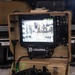 A L3Harris ground control unit (GCU) is shown open in a tan/desert-colored ruggedized carrying case. The system features a monitor displaying a live or recorded camera feed of a group of people, along with a dual-joystick controller in the lower section. This appears to be a military or defense robotic/UAS (unmanned aerial/ground system) operator control unit.