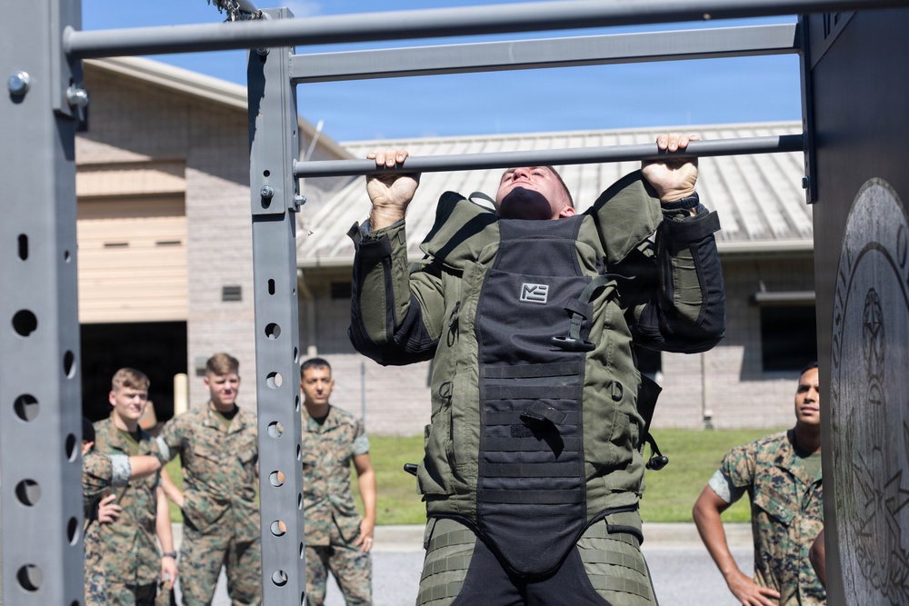A military service member in camouflage uniform and a weighted vest is performing pull-ups on an outdoor fitness rack. Several other uniformed personnel, appearing to be U.S. Marines based on their MARPAT camouflage pattern, observe in the background. The setting appears to be a military base fitness area or obstacle course.