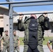 A military service member in camouflage uniform and a weighted vest is performing pull-ups on an outdoor fitness rack. Several other uniformed personnel, appearing to be U.S. Marines based on their MARPAT camouflage pattern, observe in the background. The setting appears to be a military base fitness area or obstacle course.