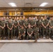 A group of approximately 30 U.S. Marines in MARPAT woodland camouflage uniforms pose for a formal group photograph inside what appears to be an ordnance or weapons training classroom. The background features shelves displaying various inert munitions, rockets, and projectiles mounted on a yellow wall. The group includes both male and female service members arranged in three rows.