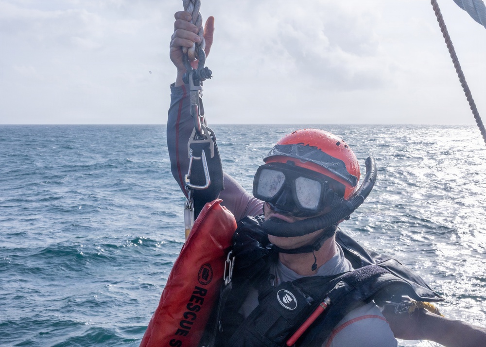 A rescue diver or water rescue personnel is photographed at sea, holding onto a rope with a carabiner clip while wearing full rescue gear. The individual is equipped with a red helmet, diving mask with snorkel, a red 'RESCUE' labeled flotation device, and a dark tactical vest. The ocean background and overcast sky suggest active maritime rescue operations or training.