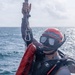 A rescue diver or water rescue personnel is photographed at sea, holding onto a rope with a carabiner clip while wearing full rescue gear. The individual is equipped with a red helmet, diving mask with snorkel, a red 'RESCUE' labeled flotation device, and a dark tactical vest. The ocean background and overcast sky suggest active maritime rescue operations or training.