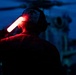 A military flight deck crew member is conducting nighttime aircraft marshalling operations using a red illuminated wand/baton. The person is wearing a helmet with markings and is positioned on what appears to be a naval vessel flight deck, with a helicopter visible in the background. The scene is dramatically lit by the red glow of the marshalling wand against a dark blue twilight sky.