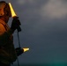 A naval flight deck crew member is photographed at dusk or dawn holding two illuminated yellow wands used for aircraft marshalling. The individual is wearing a flight deck helmet with a red anchor insignia, gloves, and a jersey, indicating a specific crew role aboard a naval vessel. The low-light conditions and glowing wands create a dramatic silhouette effect against an overcast sky.