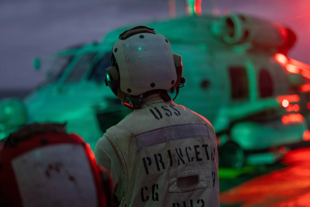 A naval crew member stands on a flight deck at dusk or night, facing a military helicopter illuminated by red and green lighting. The person's jacket is stenciled with 'USS PRINCETON CG' indicating assignment to the USS Princeton, a U.S. Navy Ticonderoga-class guided-missile cruiser. The scene depicts flight deck operations aboard a naval vessel.