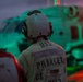 A naval crew member stands on a flight deck at dusk or night, facing a military helicopter illuminated by red and green lighting. The person's jacket is stenciled with 'USS PRINCETON CG' indicating assignment to the USS Princeton, a U.S. Navy Ticonderoga-class guided-missile cruiser. The scene depicts flight deck operations aboard a naval vessel.