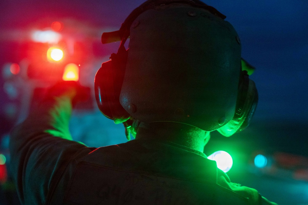 A person wearing a flight deck helmet with ear protection is photographed from behind during nighttime operations, likely on a naval vessel or airfield. They are holding a green light wand or signal light, commonly used by flight deck crew to guide aircraft. The scene is illuminated by multiple colored lights including red, orange, and green, creating a dramatic low-light atmosphere.