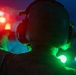 A person wearing a flight deck helmet with ear protection is photographed from behind during nighttime operations, likely on a naval vessel or airfield. They are holding a green light wand or signal light, commonly used by flight deck crew to guide aircraft. The scene is illuminated by multiple colored lights including red, orange, and green, creating a dramatic low-light atmosphere.