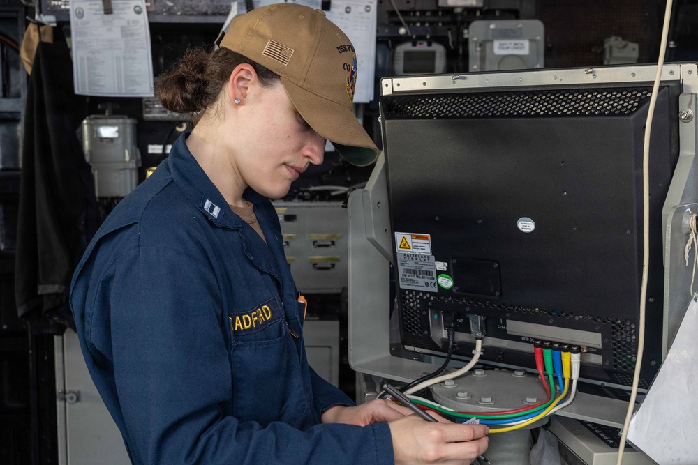A U.S. Navy officer with the name tag 'RADFORD' is working on the back of an electronic equipment unit, handling multiple colored cables. She is wearing a navy blue coverall uniform with lieutenant rank insignia and a tan baseball cap bearing a U.S. flag patch and what appears to be a ship's insignia. The setting appears to be aboard a naval vessel, likely a combat information center or technical workspace.
