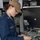 A U.S. Navy officer with the name tag 'RADFORD' is working on the back of an electronic equipment unit, handling multiple colored cables. She is wearing a navy blue coverall uniform with lieutenant rank insignia and a tan baseball cap bearing a U.S. flag patch and what appears to be a ship's insignia. The setting appears to be aboard a naval vessel, likely a combat information center or technical workspace.