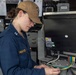 A U.S. Navy officer with the name tag 'RADFORD' is working on the back of an electronic equipment unit, handling multiple colored cables. She is wearing a navy blue coverall uniform with lieutenant rank insignia and a tan baseball cap bearing a U.S. flag patch and what appears to be a ship's insignia. The setting appears to be aboard a naval vessel, likely a combat information center or technical workspace.