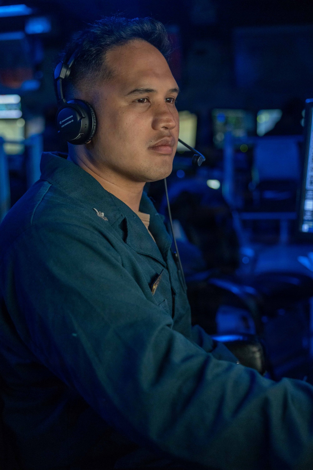 A military service member is seated at what appears to be a combat information center or operations room, wearing a Sennheiser headset with boom microphone. The environment is illuminated predominantly in blue light, consistent with a naval operations or command center setting. The individual is wearing a dark green uniform with visible insignia on the chest.
