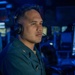 A military service member is seated at what appears to be a combat information center or operations room, wearing a Sennheiser headset with boom microphone. The environment is illuminated predominantly in blue light, consistent with a naval operations or command center setting. The individual is wearing a dark green uniform with visible insignia on the chest.