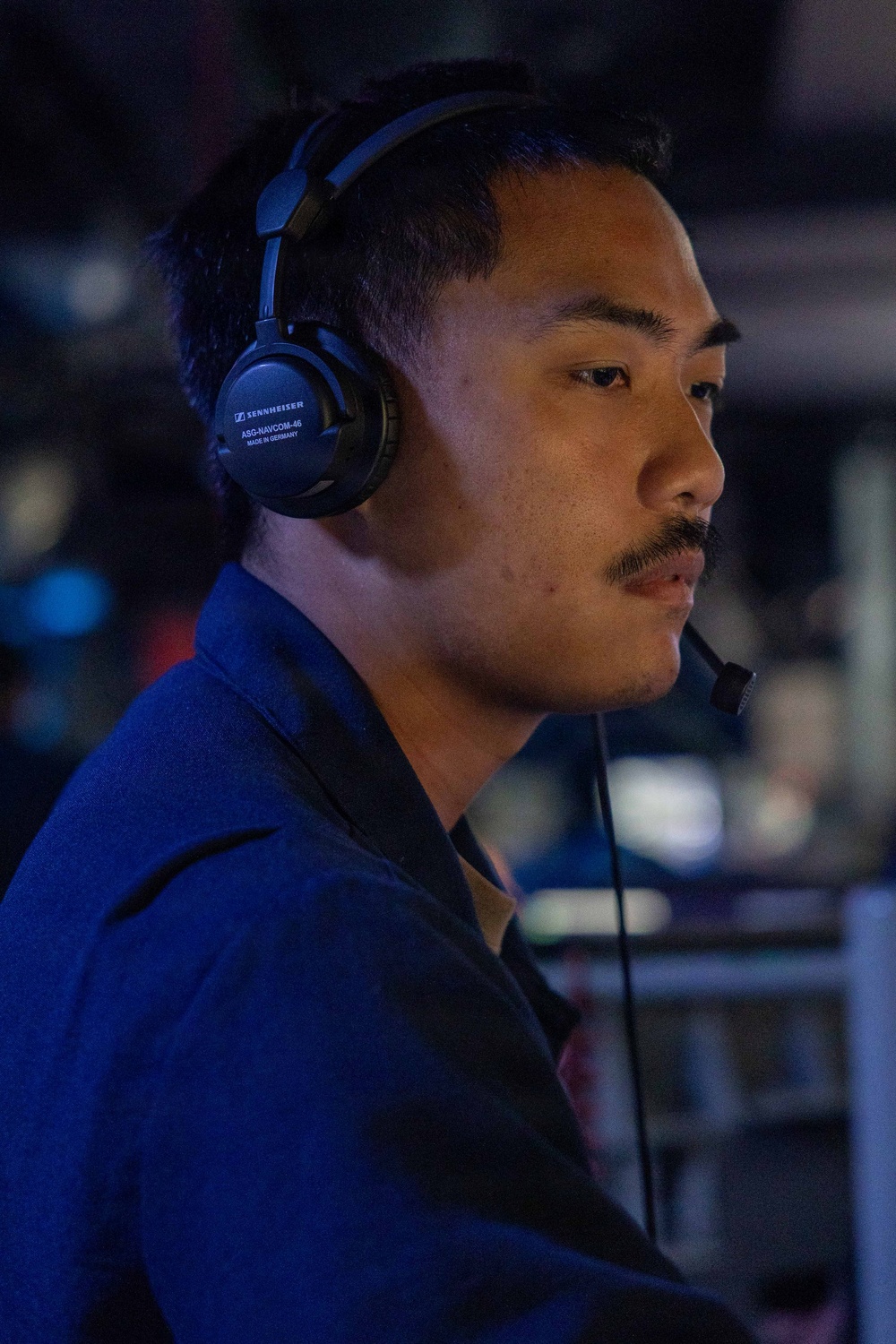 A young man wearing a Sennheiser ASG-NAVCOM-46 communications headset with a boom microphone is shown in profile view against a dark, dimly lit background. He is wearing a blue uniform or jacket and appears to be actively monitoring or communicating in what looks like a command, control, or operations center environment. Multiple screens and equipment are faintly visible in the background.