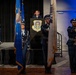 A formal military color guard ceremony is taking place in an indoor venue, with uniformed Air Force personnel presenting flags on a stage. A young woman in uniform stands at a podium bearing the insignia of the 62nd Airlift Wing, likely delivering remarks or a pledge. Four color guard members in dress blues stand at attention holding flags including the U.S. flag, Air Force flag, and a POW/MIA flag.