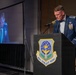 A U.S. Air Force officer in dress blues is speaking at a podium bearing the emblem of the 62nd Airlift Wing during what appears to be a formal military ceremony or banquet. The officer is wearing multiple service medals and ribbons, and is projected on a large screen behind him. The setting suggests an awards ceremony, dining-in, or official military function.