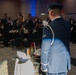 A formal military banquet or ball is taking place in a large event hall. A U.S. Air Force Honor Guard member in dress blues stands at a POW/MIA remembrance table, raising a glass in a toast. The assembled crowd of military personnel and civilians in formal attire observe the ceremony.