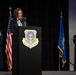 A military officer is speaking at a podium bearing the insignia of the 62d Airlift Wing during a formal ceremony. Three uniformed Air Force personnel are visible on stage, flanked by the United States flag and an Air Force flag. The setting appears to be an indoor auditorium or event hall with stage lighting.