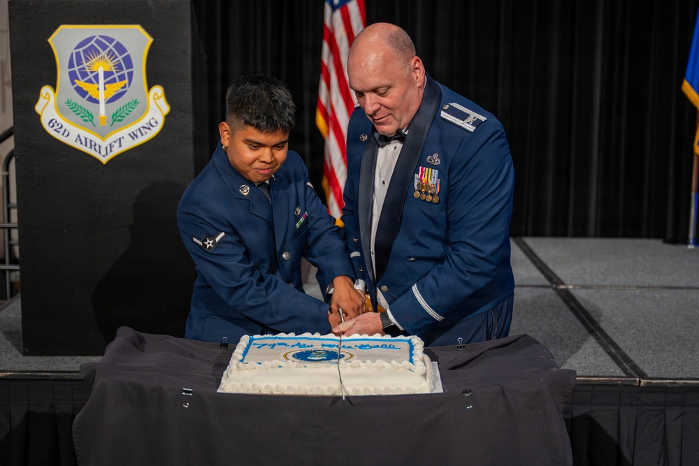 Two U.S. Air Force personnel in dress blues are cutting a ceremonial cake at what appears to be an Air Force birthday ball or formal military celebration. A junior enlisted Airman and a senior officer (identifiable by rank insignia and medals) are jointly cutting the cake using a ceremonial saber. The event is associated with the 62d Airlift Wing, as indicated by the emblem visible on the podium in the background.
