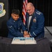 Two U.S. Air Force personnel in dress blues are cutting a ceremonial cake at what appears to be an Air Force birthday ball or formal military celebration. A junior enlisted Airman and a senior officer (identifiable by rank insignia and medals) are jointly cutting the cake using a ceremonial saber. The event is associated with the 62d Airlift Wing, as indicated by the emblem visible on the podium in the background.