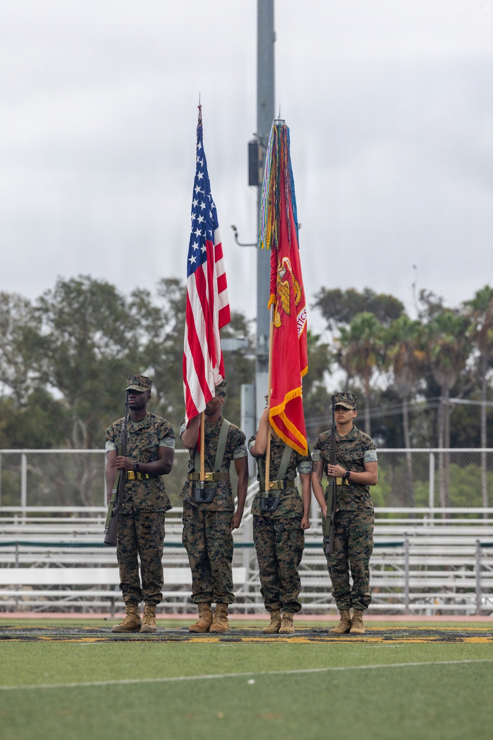 A U.S. Marine Corps Color Guard stands at attention on a football field, presenting the United States national flag and the Marine Corps flag with battle streamers. The four Marines are in full dress woodland MARPAT camouflage uniforms with rifles and ceremonial equipment. The ceremony appears to be taking place at an outdoor athletic stadium with bleachers visible in the background.