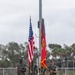 A U.S. Marine Corps Color Guard stands at attention on a football field, presenting the United States national flag and the Marine Corps flag with battle streamers. The four Marines are in full dress woodland MARPAT camouflage uniforms with rifles and ceremonial equipment. The ceremony appears to be taking place at an outdoor athletic stadium with bleachers visible in the background.