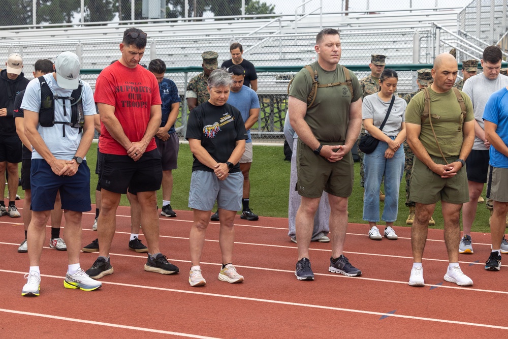 A group of civilians and military personnel stand on a running track with heads bowed and hands clasped, appearing to observe a moment of silence or prayer. The setting is an athletic facility with bleachers visible in the background. Some participants wear military uniforms while others are in civilian athletic attire, suggesting a joint commemorative or memorial event.