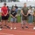 Strength in Every Step: Laps For Life event supports suicide awareness A group of civilians and military personnel stand on a running track with heads bowed and hands clasped, appearing to observe a moment of silence or prayer. The setting is an athletic facility with bleachers visible in the background. Some participants wear military uniforms while others are in civilian athletic attire, suggesting a joint commemorative or memorial event.