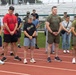 A group of civilians and military personnel stand on a running track with heads bowed and hands clasped, appearing to observe a moment of silence or prayer. The setting is an athletic facility with bleachers visible in the background. Some participants wear military uniforms while others are in civilian athletic attire, suggesting a joint commemorative or memorial event.