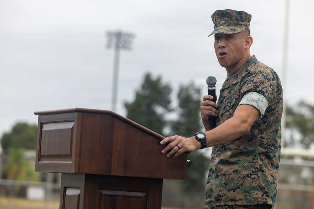 A U.S. Marine Corps service member in MARPAT digital camouflage uniform is speaking at a wooden podium while holding a handheld microphone. The setting appears to be an outdoor military ceremony or event, likely on a military installation. The speaker is wearing a cover (cap) and has a watch and ring visible on his hands.