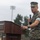 Strength in Every Step: Laps For Life event supports suicide awareness A U.S. Marine Corps service member in MARPAT digital camouflage uniform is speaking at a wooden podium while holding a handheld microphone. The setting appears to be an outdoor military ceremony or event, likely on a military installation. The speaker is wearing a cover (cap) and has a watch and ring visible on his hands.