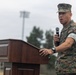 A U.S. Marine Corps service member in MARPAT digital camouflage uniform is speaking at a wooden podium while holding a handheld microphone. The setting appears to be an outdoor military ceremony or event, likely on a military installation. The speaker is wearing a cover (cap) and has a watch and ring visible on his hands.