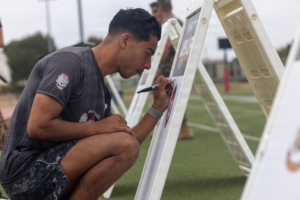 A young man crouches down and writes or signs on a whiteboard easel on what appears to be an athletic field. He is wearing a gray graphic t-shirt featuring a Día de los Muertos skull design and camouflage shorts. Several similar easels are visible in the background, suggesting a group activity or event.