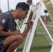 A young man crouches down and writes or signs on a whiteboard easel on what appears to be an athletic field. He is wearing a gray graphic t-shirt featuring a Día de los Muertos skull design and camouflage shorts. Several similar easels are visible in the background, suggesting a group activity or event.