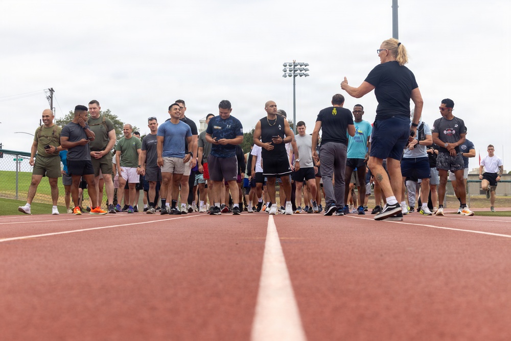 A large group of people dressed in athletic/casual workout attire are gathered on a red running track, appearing to receive instructions or a briefing from a person standing with their back to the camera. The setting appears to be an outdoor sports facility with stadium lighting visible in the background. The gathering suggests a organized fitness event, race, or group workout session.