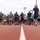 Strength in Every Step: Laps For Life event supports suicide awareness A large group of people dressed in athletic/casual workout attire are gathered on a red running track, appearing to receive instructions or a briefing from a person standing with their back to the camera. The setting appears to be an outdoor sports facility with stadium lighting visible in the background. The gathering suggests a organized fitness event, race, or group workout session.