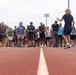 A large group of people dressed in athletic/casual workout attire are gathered on a red running track, appearing to receive instructions or a briefing from a person standing with their back to the camera. The setting appears to be an outdoor sports facility with stadium lighting visible in the background. The gathering suggests a organized fitness event, race, or group workout session.