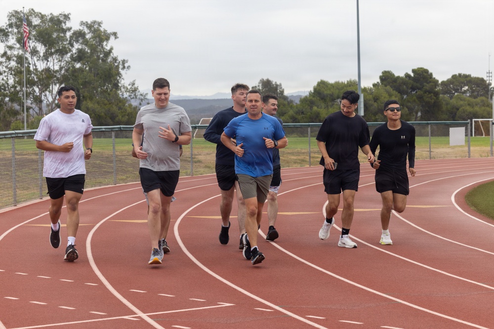 A group of seven men are running together on a red outdoor athletic track under overcast skies. They are dressed in casual athletic wear including t-shirts and shorts, suggesting a group fitness or physical training session. An American flag is visible in the background along with trees, hills, and stadium lighting poles.