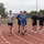 Strength in Every Step: Laps For Life event supports suicide awareness A group of seven men are running together on a red outdoor athletic track under overcast skies. They are dressed in casual athletic wear including t-shirts and shorts, suggesting a group fitness or physical training session. An American flag is visible in the background along with trees, hills, and stadium lighting poles.