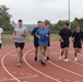 A group of seven men are running together on a red outdoor athletic track under overcast skies. They are dressed in casual athletic wear including t-shirts and shorts, suggesting a group fitness or physical training session. An American flag is visible in the background along with trees, hills, and stadium lighting poles.