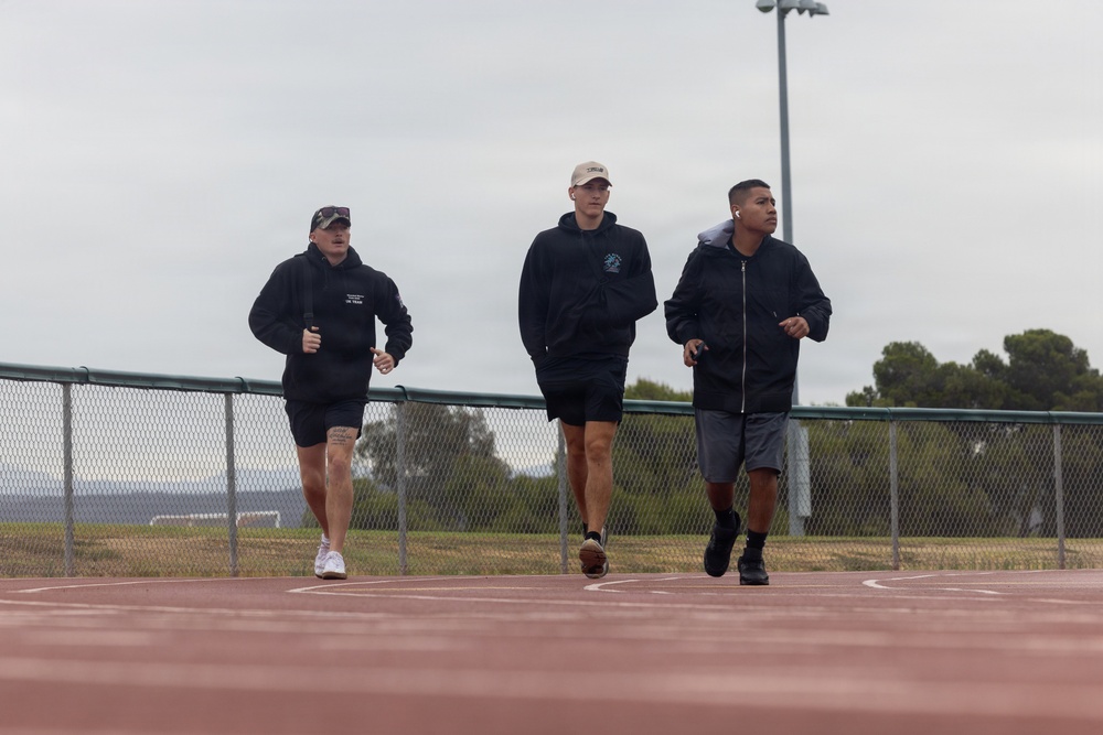 Three individuals are jogging or walking on an outdoor running track, all dressed in dark athletic wear. The setting appears to be a sports facility with a chain-link fence, light poles, and trees visible in the background under an overcast sky. The group appears to be engaged in a casual fitness activity together.